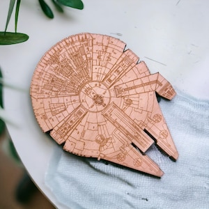May include: A wooden coaster in the shape of the Millennium Falcon spaceship. The coaster is light brown with detailed engraved lines depicting the ship's exterior. The coaster is resting on a light blue cloth.