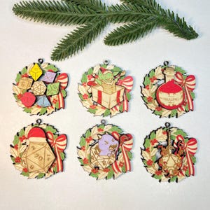 Op de afbeelding: Zes houten kerstversieringen met een kransontwerp. Elke versiering heeft een ander fantasiethema, waaronder dobbelstenen, een goblin in een geschenkdoos, een drankfles en een gitaar. Rode en groene accenten worden door het hele ontwerp gebruikt.