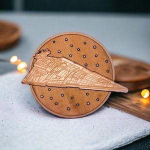 May include: A wooden coaster featuring a detailed laser-cut image of a Star Destroyer spaceship. The coaster is round with a dark brown finish and small star cutouts. The spaceship is light brown.