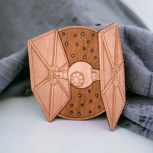 May include: A wooden TIE fighter, a spacecraft from a science fiction franchise, is displayed. The spacecraft is light brown with detailed laser-cut lines. The background features a starry night design. The wings are angled outward.