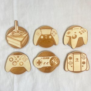 Könnte beinhalten: Sechs Untersetzer aus Holz mit verschiedenen Videospiel-Controller-Designs. Enthält einen Atari-Joystick, PlayStation, Nintendo 64, Xbox, Super Nintendo und Nintendo Switch Controller. Jeder Untersetzer ist aus hellem Holz mit gravierten Details.