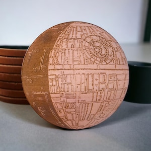 May include: A round wooden coaster with a detailed engraved design of the Death Star from Star Wars. The coaster is light brown with a detailed pattern. Other coasters and a dark green mug are in the background.