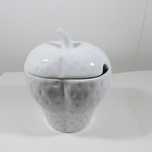 May include: A white ceramic strawberry-shaped container with a lid and a small spoon opening. The container has a textured surface and a stem-like handle on the lid. The design is simple and elegant.