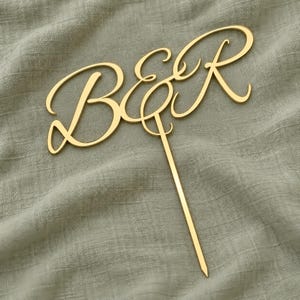 May include: A gold-colored cake topper featuring the letters B and R connected by an ampersand in a flowing, cursive script font. The design includes a single pointed stake extending downward from the center of the ampersand for insertion.