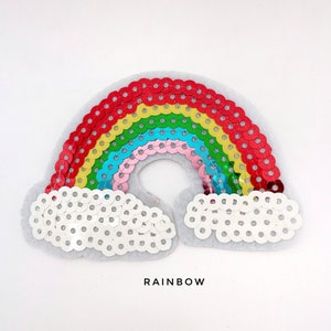 Può includere: Un cerotto di paillettes a forma di arcobaleno con un retro in feltro bianco. L'arcobaleno è fatto di paillettes rosse, arancioni, gialle, verdi, blu, rosa e viola. Il cerotto ha due nuvole bianche su entrambi i lati dell'arcobaleno. Il testo "RAINBOW" è stampato sotto il cerotto.