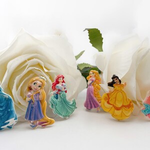 Op de afbeelding: Zes kleine plastic figuurtjes van Disney-prinsessen, waaronder Assepoester, Rapunzel, Ariel, Belle, Aurora en Sneeuwwitje. De figuurtjes zijn gerangschikt op een witte achtergrond met een witte roos.