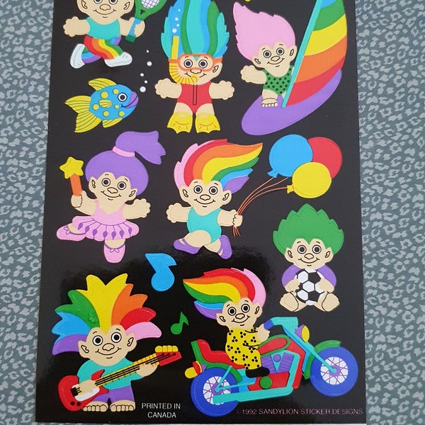 Può includere: Un foglio di adesivi colorati con vari personaggi troll. I troll sono raffigurati in diverse attività, tra cui giocare a tennis, guidare una motocicletta e tenere palloncini. Gli adesivi sono stampati su uno sfondo nero e hanno una finitura lucida.