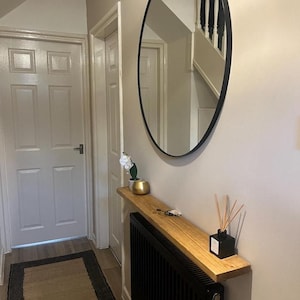 May include: A narrow wooden shelf is mounted above a black radiator. The shelf is decorated with a small potted plant, a gold decorative object, and a diffuser with reeds. A large round mirror with a black frame hangs on the wall above the shelf.