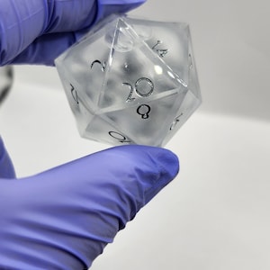 Puede incluir: Un dado transparente de veinte caras con números negros. El dado se sostiene en una mano enguantada. Los números son claramente visibles, con el número 20 en un lugar destacado. Este dado poliédrico es probablemente para juegos de mesa.