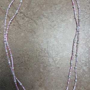 May include: A delicate, double-strand necklace featuring small pink and white beads, interspersed with larger, pearlescent beads. The necklace is secured with a gold-colored clasp. The overall design is feminine and elegant.