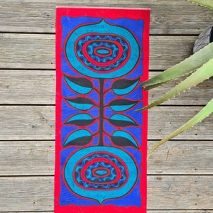 Op de afbeelding: Een rechthoekig kunstwerk met een opvallend bloemmotief. Het heeft een rode rand, een blauwe achtergrond en twee grote, gestileerde turquoise bloemen. Zwarte bladeren en stelen verbinden de bloemen. Het kunstwerk staat op een houten oppervlak.