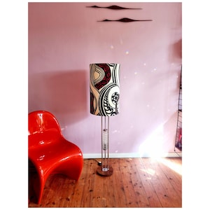 May include: A floor lamp with a white, black, and red patterned lampshade. The lamp has a chrome base and a clear glass cylinder in the middle. The lamp is in a room with a red chair and a pink wall.