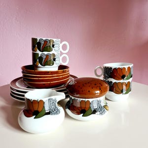 Op de afbeelding: Een vintage set van bruine en witte, met bloemen geprinte, keramische theekopjes, schoteltjes, een roomkan en een suikerpot. De set heeft een bruin en wit bloemenpatroon met een bruine rand.