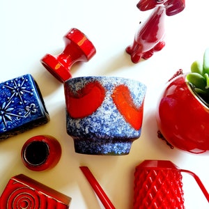 Pode incluir: Uma coleção de vasos e objetos decorativos de cerâmica vermelha e azul. Os vasos apresentam um esmalte azul e branco salpicado com detalhes vermelhos. Os objetos decorativos incluem uma figura de cão, um regador e um pequeno porta-velas.