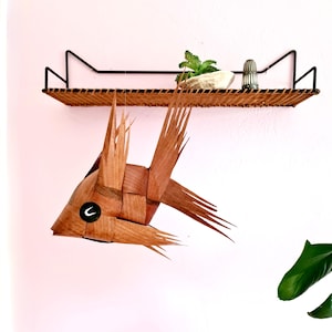May include: A woven rattan shelf with a black metal frame, holding a small plant, a rock, and a silver object. Below, a decorative fish made of woven wood hangs from the shelf, featuring a black and white eye detail. The background is a pale pink wall.