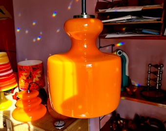 May include: A vibrant orange glass pendant light fixture with a rounded, hourglass shape. The light hangs from a black cord and a silver-toned metal cap. The fixture is a retro design, likely from the mid-century era.