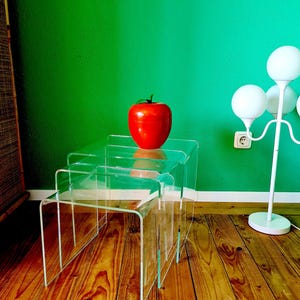 May include: A set of three clear acrylic nesting tables. A large red apple with a green stem sits on the top table. The tables are on a wooden floor, with a green wall in the background.