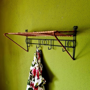 Könnte beinhalten: Ein an der Wand montierter Kleiderständer mit Ablage und Haken. Der Ständer ist aus schwarzem Metall mit einem gewebten braunen Regal und Akzenten. Ein weißes Hemd mit rotem und grünem Blumenmuster hängt an einem der Haken an einer grünen Wand.