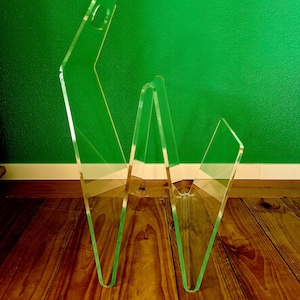 Könnte beinhalten: Ein durchsichtiges Acryl-Zeitschriftenregal mit einem einzigartigen, kantigen Design. Das Regal hat eine Reihe von verbundenen, V-förmigen Paneelen, die mehrere Ablagefächer bilden. Das transparente Material ermöglicht die Sichtbarkeit des Inhalts. Das Regal steht auf einem Holzboden vor einer grünen Wand.