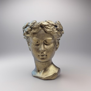 Op de afbeelding: Een goudkleurige buste van een vrouwenhoofd met gesloten ogen en een lauwerkrans. De sculptuur heeft een getextureerd oppervlak en een matte afwerking. Het hoofd staat tegen een zachte, neutrale achtergrond.