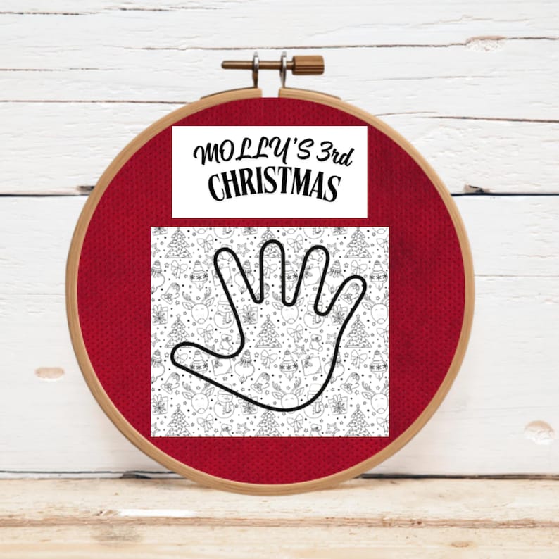A red embroidery hoop with a white rectangle featuring the text "MOLLY'S 3rd CHRISTMAS". Below is a patterned design with a hand outline and Christmas-themed illustrations in black and white.