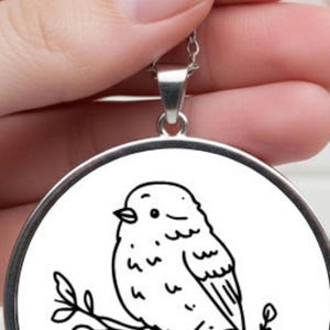 A silver pendant necklace with a circular pendant. The pendant displays a black and white line drawing of a bird on a branch. The necklace is held up by a hand. The bird is drawn in a simple style.