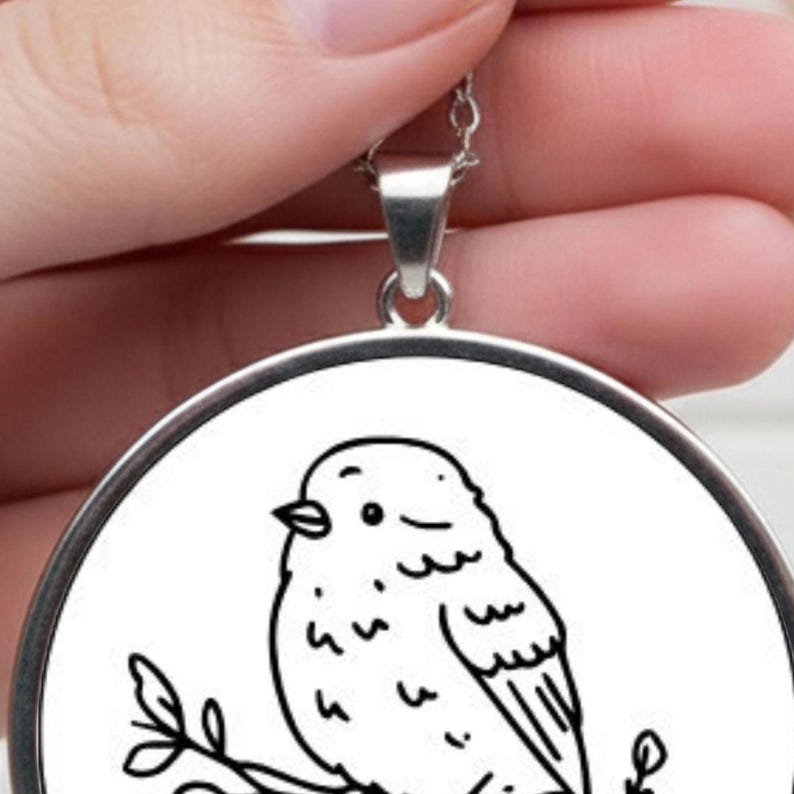 A silver pendant necklace with a circular pendant. The pendant displays a black and white line drawing of a bird on a branch. The necklace is held up by a hand. The bird is drawn in a simple style.