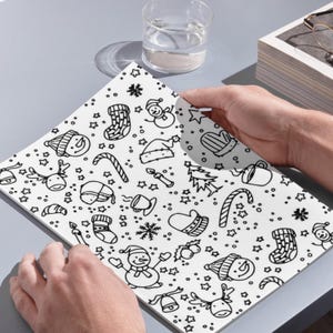 In image, a hand holds open a white book featuring a detailed pattern of hand-drawn Christmas doodles. Small illustrations like smiling snowmen, decorated Christmas trees, presents, candy canes, mistletoe, and stars are scattered across the pages in black line art.

Above the book, a clear glass of water sits next to a small stack of white books. The objects rest on a grey surface and catch natural daylight. This overhead perspective captures the open book as the central focus.