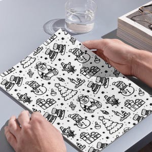 In image, a hand holds open a white book featuring a detailed pattern of hand-drawn Christmas doodles. Small illustrations like smiling snowmen, stylized Christmas trees, wrapped presents, teddy bears, candles, and snowflakes are scattered across the pages in black line art.

Above the book, a clear glass of water sits next to a small stack of white books. The objects rest on a grey surface and catch natural daylight. This overhead perspective captures the open book as the central focus.