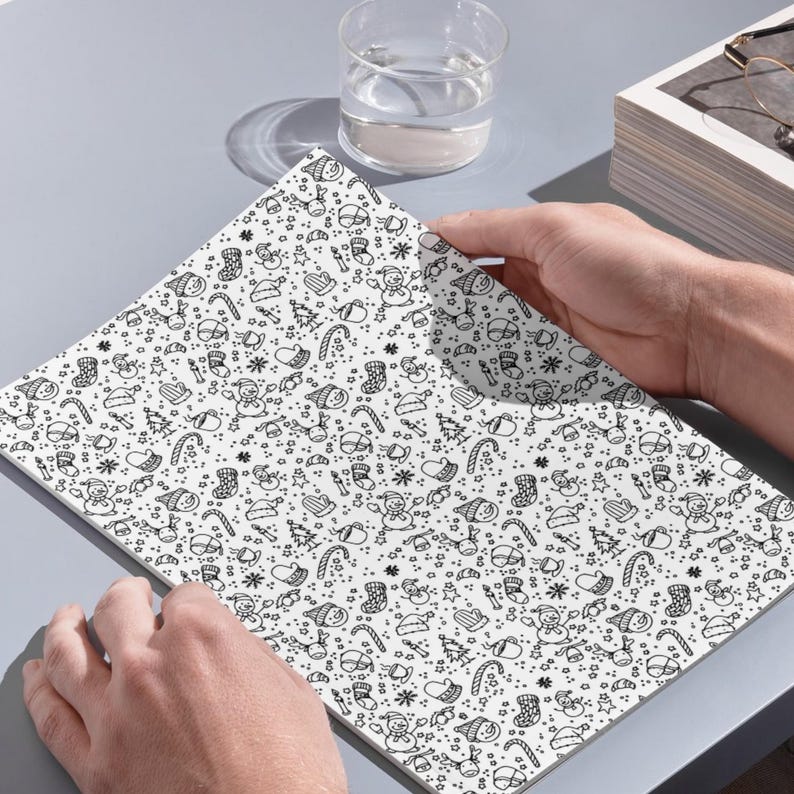 In image, a hand holds open a white book featuring a detailed pattern of hand-drawn Christmas doodles. Small illustrations like smiling snowmen, decorated Christmas trees, presents, candy canes, mistletoe, and stars are scattered across the pages in black line art.

Above the book, a clear glass of water sits next to a small stack of white books. The objects rest on a grey surface and catch natural daylight. This overhead perspective captures the open book as the central focus.