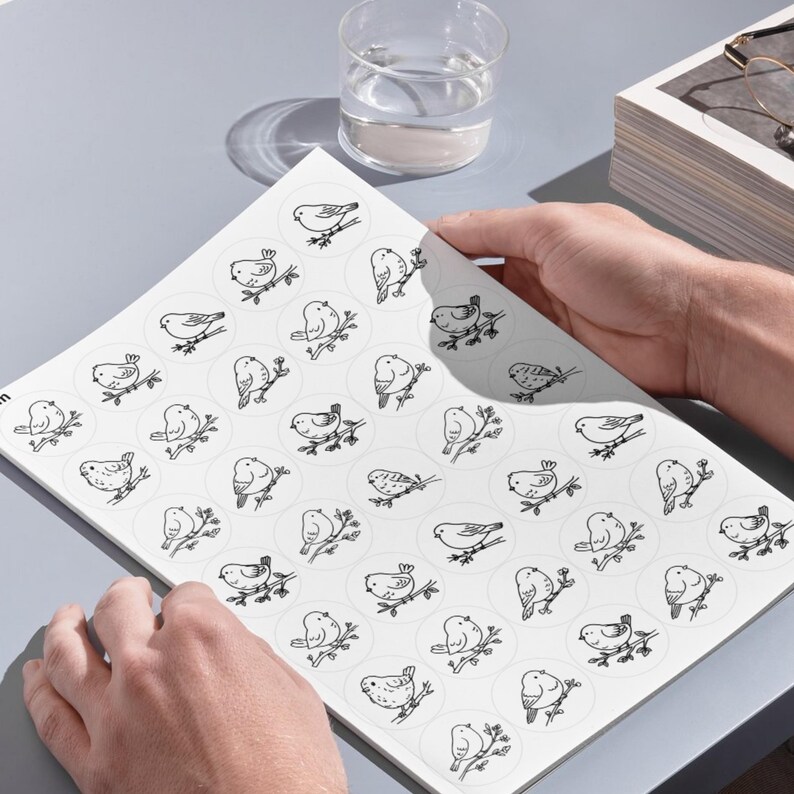 A high-angle photograph showing a person's hands holding a large white sheet on a smooth, grey desk surface. The sheet, referenced as image, contains several rows of large circular templates, each displaying a detailed black line-art illustration of a bird perched on a branch. In the upper portion of the background, a clear glass of water, a stack of books, and a pair of eyeglasses are arranged under clean, bright lighting that casts soft shadows across the setup.