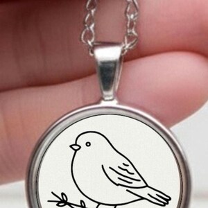 Silver necklace with a circular pendant displaying a black and white drawing of a bird on a branch. The bird illustration is minimalist. The necklace has a chain.