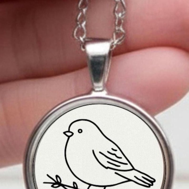 Silver necklace with a circular pendant displaying a black and white drawing of a bird on a branch. The bird illustration is minimalist. The necklace has a chain.