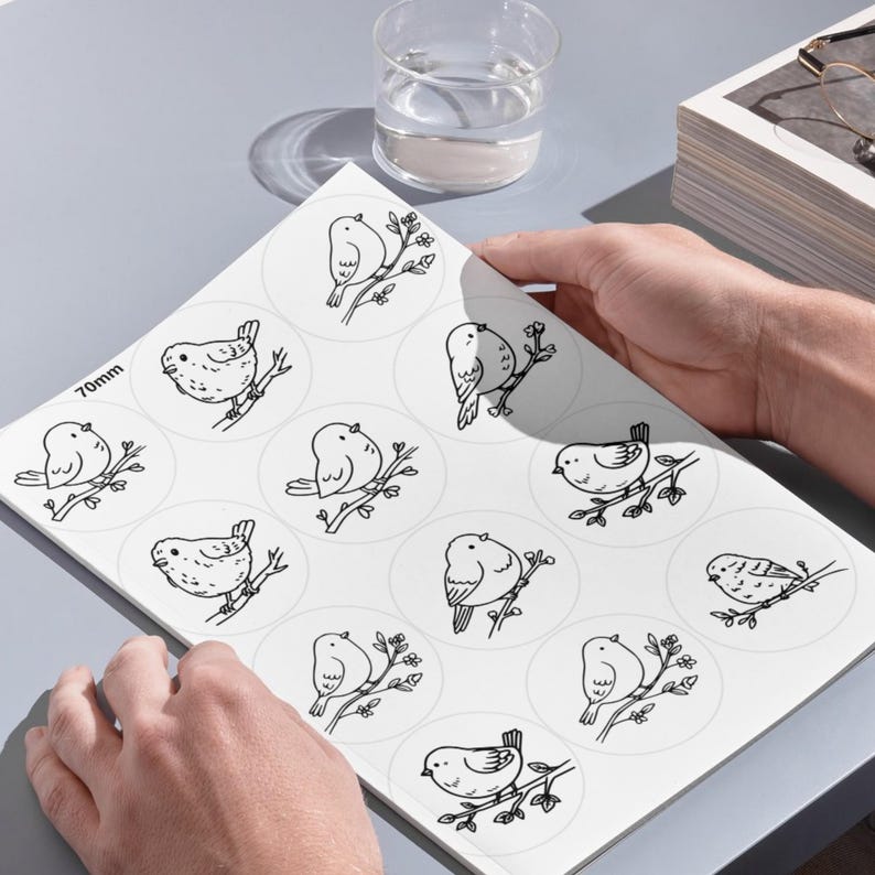 A high-angle photograph showing a person's hands holding a large white sheet on a smooth, grey desk surface. The sheet, referenced as image, contains several rows of large circular templates, each displaying a detailed black line-art illustration of a bird perched on a branch. In the upper portion of the background, a clear glass of water, a stack of books, and a pair of eyeglasses are arranged under clean, bright lighting that casts soft shadows across the setup.