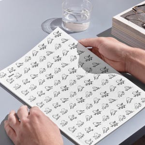 A person's hands holding a large white sheet in image_a8df2e.jpg printed with dozens of 25mm circular stick-and-stitch embroidery stabilizers on a grey desk. Each circle features a delicate, black line-art illustration of a bird perched on a branch. A glass of water, a pair of glasses, and a stack of books are visible in the background under clean lighting.