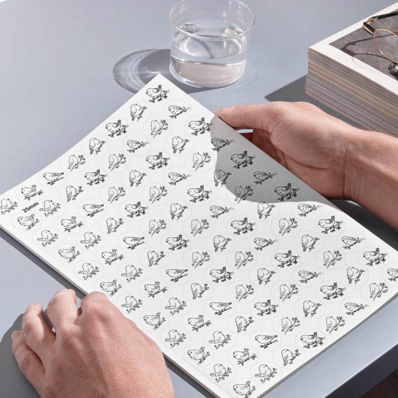 A person's hands holding a large white sheet in image_a8df2e.jpg printed with dozens of 25mm circular stick-and-stitch embroidery stabilizers on a grey desk. Each circle features a delicate, black line-art illustration of a bird perched on a branch. A glass of water, a pair of glasses, and a stack of books are visible in the background under clean lighting.