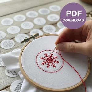 May include: A wooden embroidery hoop holds white fabric with a red snowflake design being stitched. A needle and red thread are used to create the embroidery. The image includes a purple circle with the text "PDF DOWNLOAD" and several small, circular designs.