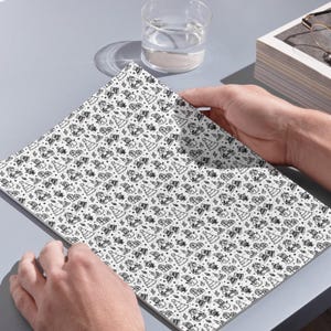 In image, a hand holds open a white book featuring a detailed pattern of hand-drawn Christmas doodles. Small illustrations like smiling snowmen, stylized Christmas trees, wrapped presents, teddy bears, candles, and snowflakes are scattered across the pages in black line art.

Above the book, a clear glass of water sits next to a small stack of white books. The objects rest on a grey surface and catch natural daylight. This overhead perspective captures the open book as the central focus.
