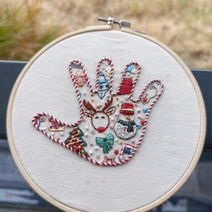 Peut inclure: Une broderie en forme de main sur tissu blanc, fixée dans un tambour en bois. Le contour est réalisé avec un fil torsadé rouge et blanc. L'intérieur présente des motifs de Noël colorés, dont un renne, un bonhomme de neige et un sapin.