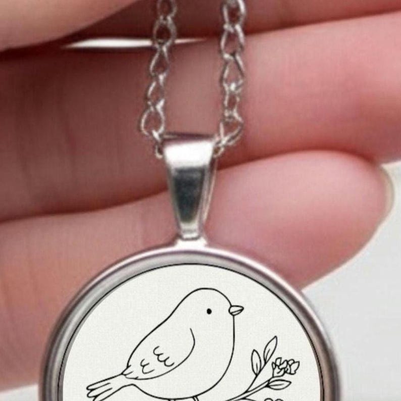 Silver necklace with a circular pendant. The pendant displays a black and white drawing of a bird on a branch with flowers. The pendant is about 2.5 cm in diameter, hanging from a fine chain.