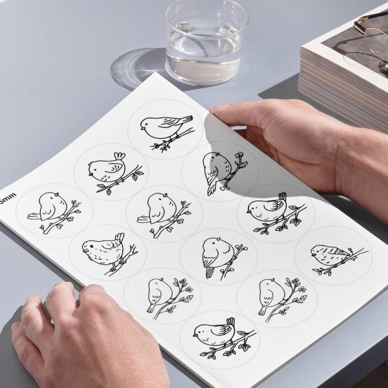 A high-angle photograph showing a person's hands holding a large white sheet on a smooth, grey desk surface. The sheet, referenced as image, contains several rows of large circular templates, each displaying a detailed black line-art illustration of a bird perched on a branch. In the upper portion of the background, a clear glass of water, a stack of books, and a pair of eyeglasses are arranged under clean, bright lighting that casts soft shadows across the setup.