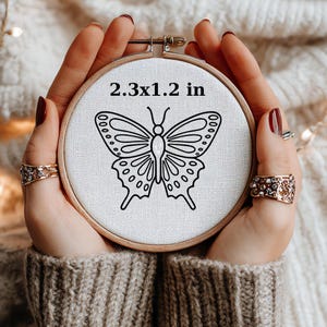 May include: A wooden embroidery hoop with a butterfly design. The text "2.3x1.2 in" is printed on the fabric. The butterfly is black and has detailed wing patterns. The hoop is held by hands wearing rings.