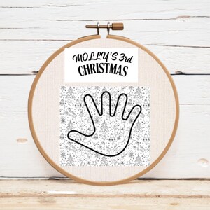 A wooden embroidery hoop holding a fabric panel with a hand outline and Christmas-themed patterns. The text "MOLLY'S 3rd CHRISTMAS" is printed at the top of the panel.