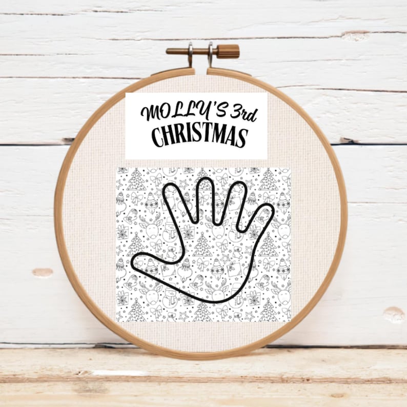 A wooden embroidery hoop holding a fabric panel with a hand outline and Christmas-themed patterns. The text "MOLLY'S 3rd CHRISTMAS" is printed at the top of the panel.
