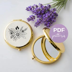 Może przedstawiać: Dwa okrągłe, złote lusterka kieszonkowe. Jedno jest zamknięte, z białą tkaniną na wierzchu ozdobioną czarną grafiką kwiatową. Drugie lusterko jest otwarte, ukazując powierzchnię odbijającą. Obok leży gałązka fioletowej lawendy.