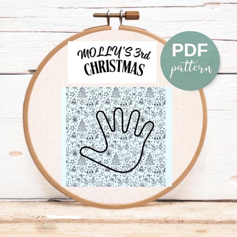 An embroidery hoop on white wood showcases a Christmas embroidery pattern. Text reads "MOLLY'S 3rd CHRISTMAS" and "PDF pattern." Below, a black outline of a handprint sits over a light blue background filled with small holiday doodles like trees, reindeer, and snowmen.