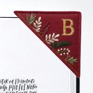 May include: A triangular red felt corner bookmark featuring embroidered botanical details in green, white, and red, along with a prominent gold-stitched letter B. The bookmark is positioned on the corner of a white page with visible black text.