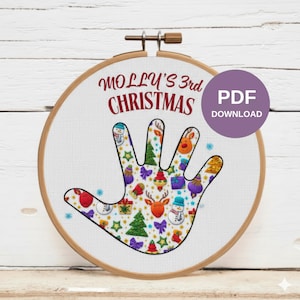 Puede incluir: Un bastidor de bordado con un diseño en forma de mano lleno de ilustraciones navideñas. El texto "MOLLY'S 3rd CHRISTMAS" está arqueado sobre la mano, con un botón "PDF DOWNLOAD" a la derecha. El bastidor está sobre un fondo de madera blanca.