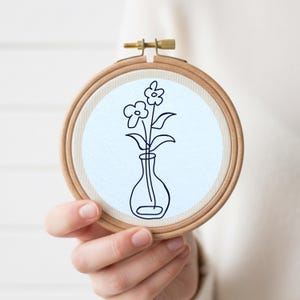 May include: A floral embroidery hoop featuring a minimalist line drawing of flowers in a vase. The design is in black against a light blue background, set within a wooden hoop. The hoop is held by a person.