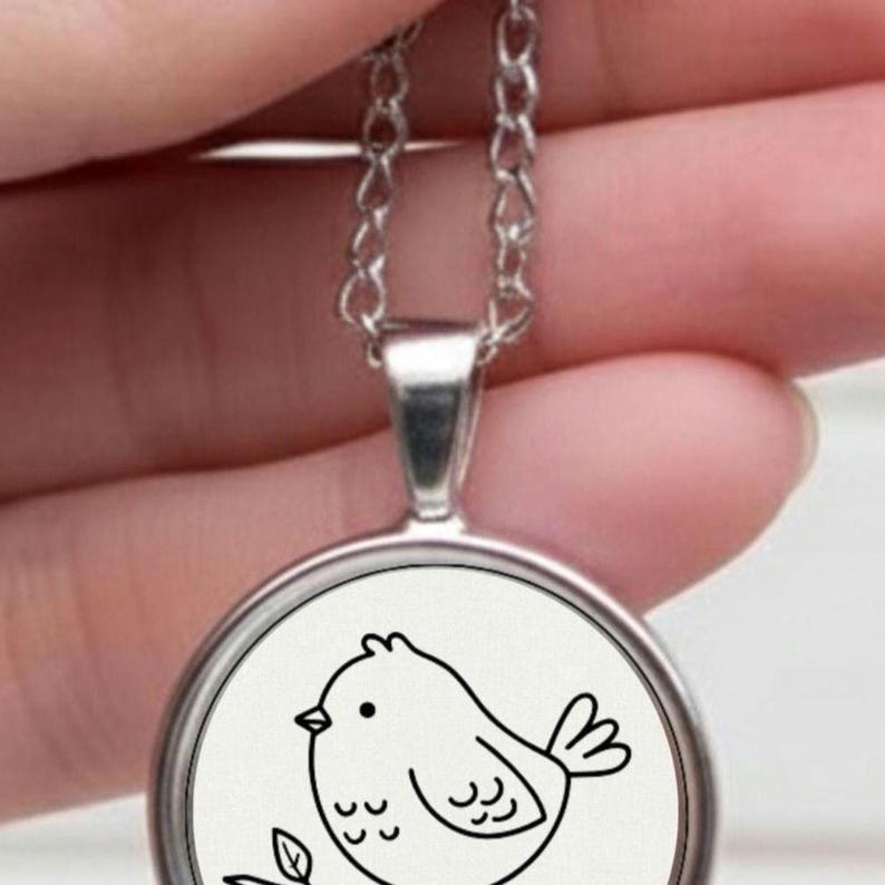 Silver necklace with a circular pendant. The pendant displays a black and white drawing of a bird on a branch with leaves. The bird is depicted in a simple, cartoon style. The chain is fine and attached to a silver bail.